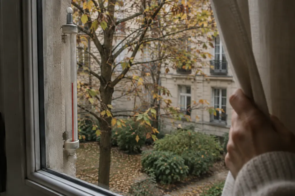 Une main ouvre un rideau près d’un thermomètre flou donnant sur une cour d’automne.